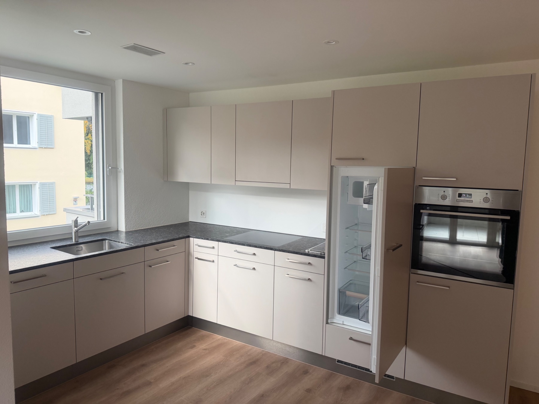 Helle Küche mit Griffschrankfronten, Granit-/Schiefer-Arbeitsfläche sowie eingebautem Backofen und Kühlschrank. Sichtbar ist der gereinigte Küchenbereich mit Bodenbelag.